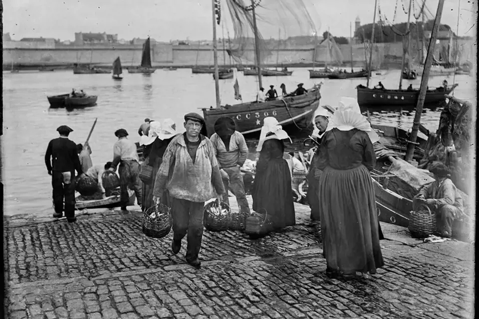 Photographie ancienne de pêcheurs en béret et vareuse déchargeant leur pêche dans des paniers d'osier sur le quai de Concarneau